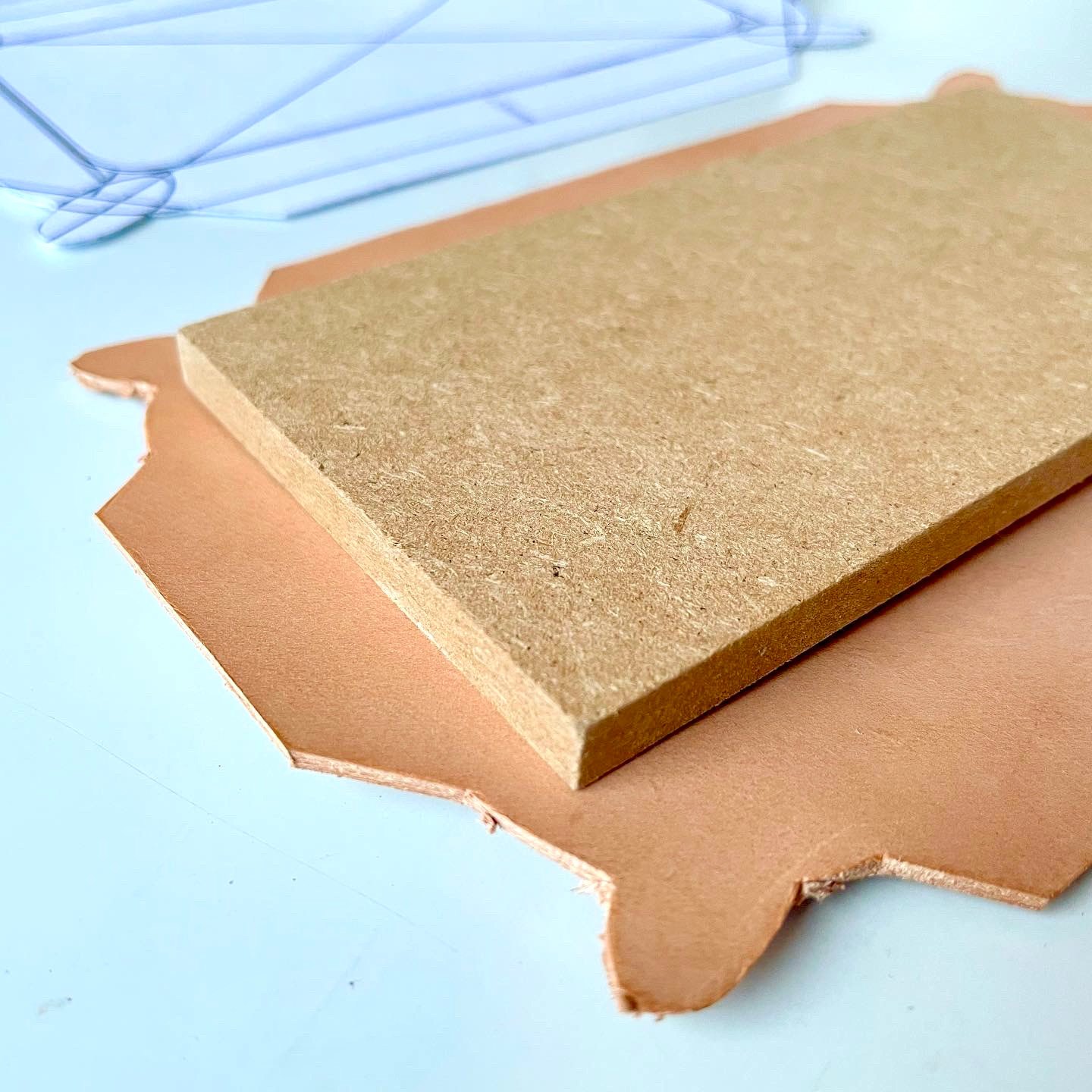 Ein Stück pflanzlich gegerbtes Leder mit noch unbeschnittenen Kanten unter einem Block aus Faserplatte auf einem weißen Tisch, dahinter Papierschablonen