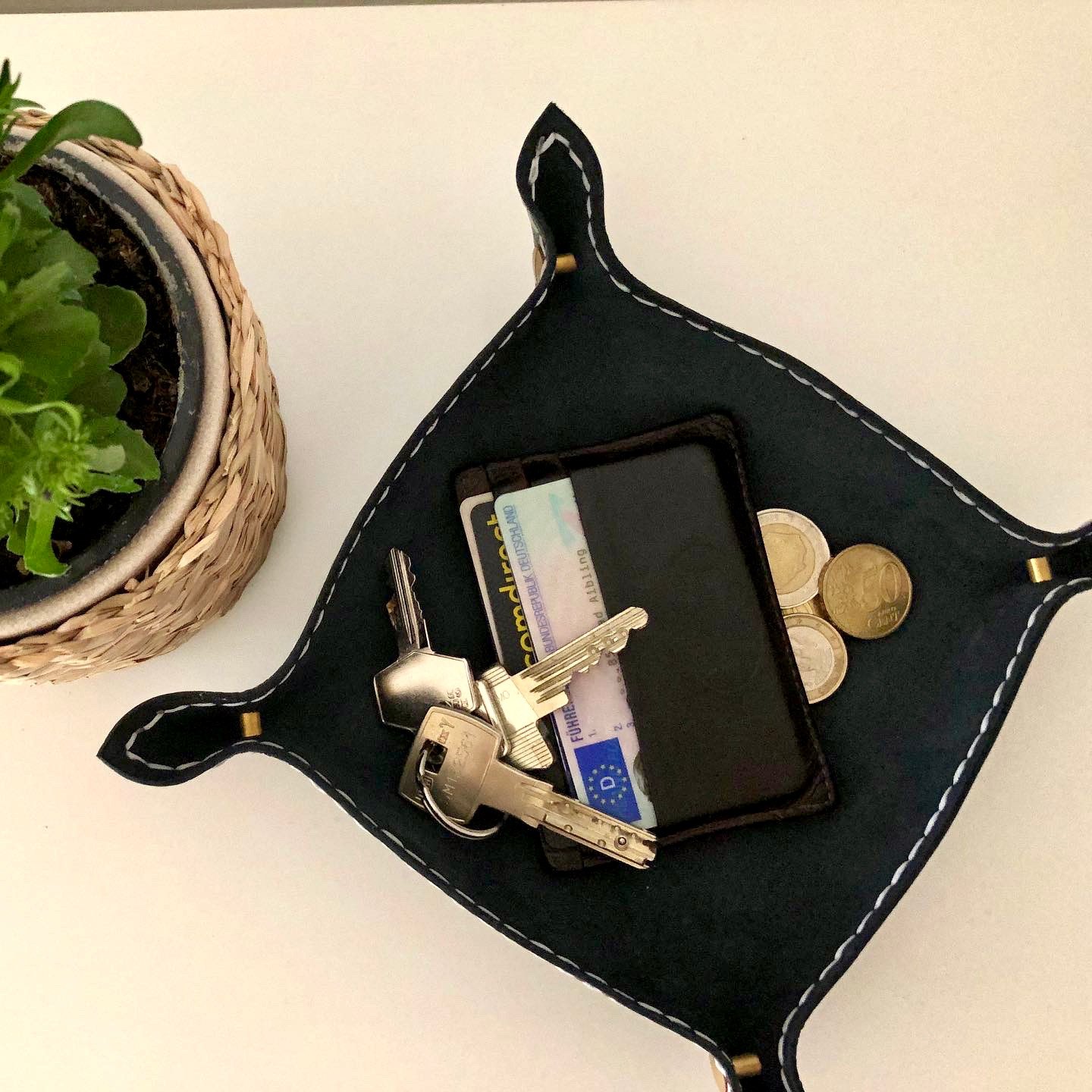 Black valet tray by 1985 Studio in chrome-tanned leather from above, its four corners fastened with brass Chicago screws and white stitching around the edge, holding keys, a card wallet and coins beside a potted plant