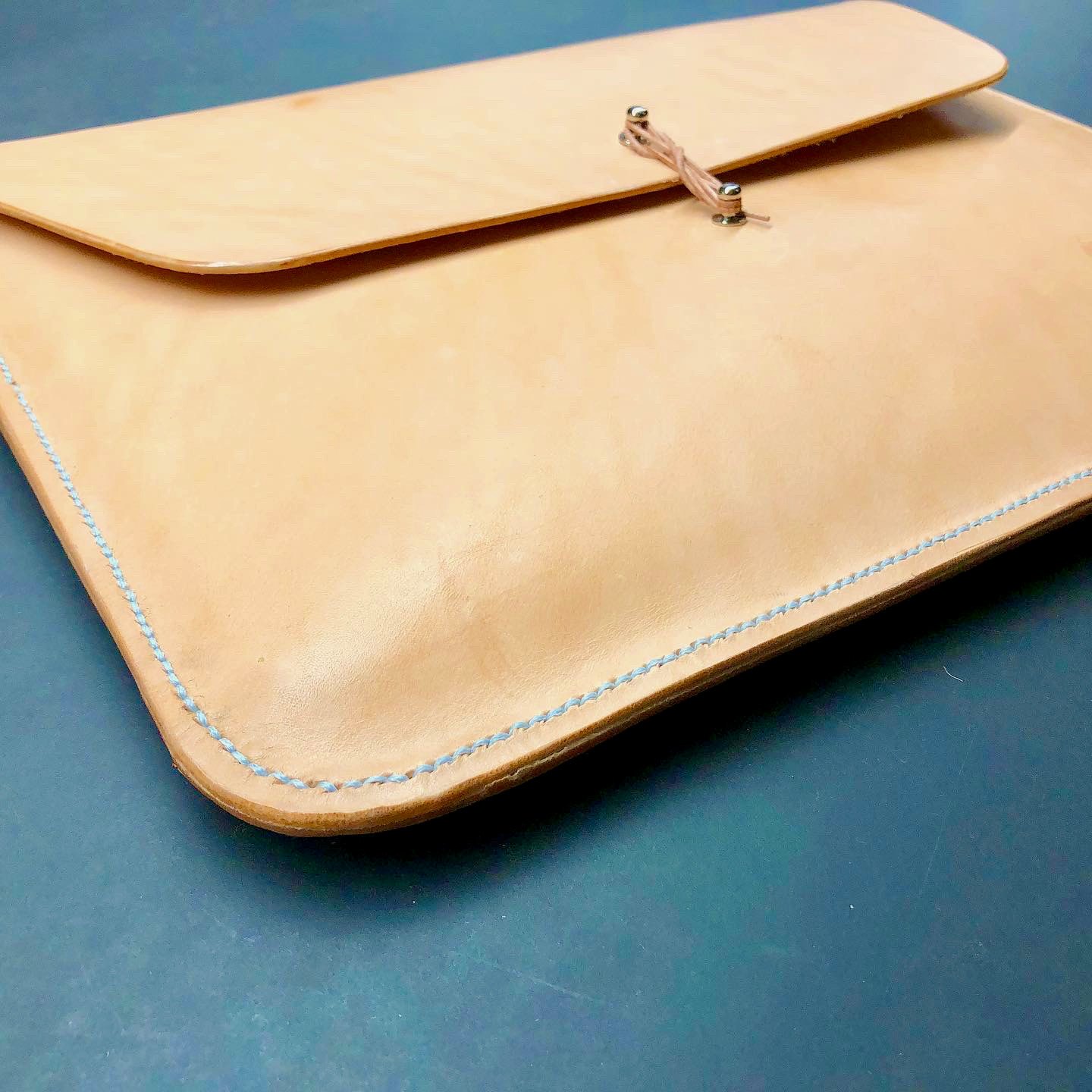 Envelope laptop case by 1985 Studio in vegetable-tanned leather at an angle, its flap and string closure, the edge stitched in pale blue thread