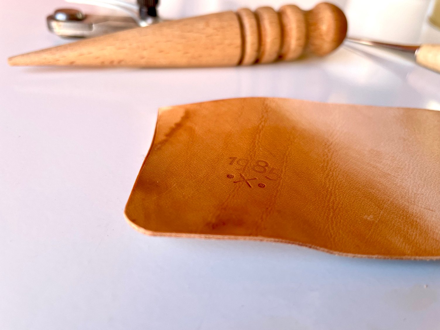 Ein Stück pflanzlich gegerbtes Leder, mit Öl und Bienenwachs nachgedunkelt und mit der Marke 1985 Studio geprägt, vor einem hölzernen Kantenglätter