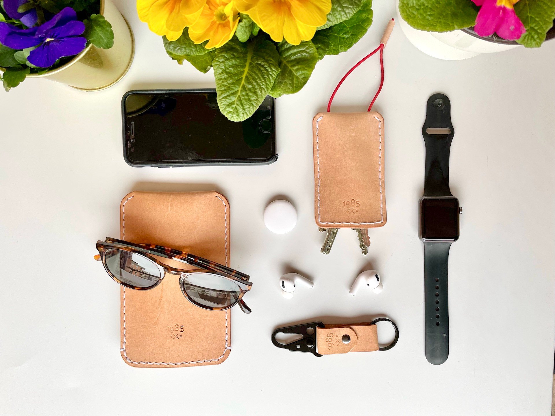 Handgemachte Lederwaren von 1985 Studio auf einem weißen Tisch unter Frühlingsblumen: ein Sonnenbrillen-Etui unter einer Schildpatt-Sonnenbrille, ein Schlüsseletui mit rotem Zugband und zwei Schlüsseln und ein Schlüsselanhänger mit schwarzem Karabiner, daneben iPhone, AirTag, AirPods und Apple Watch