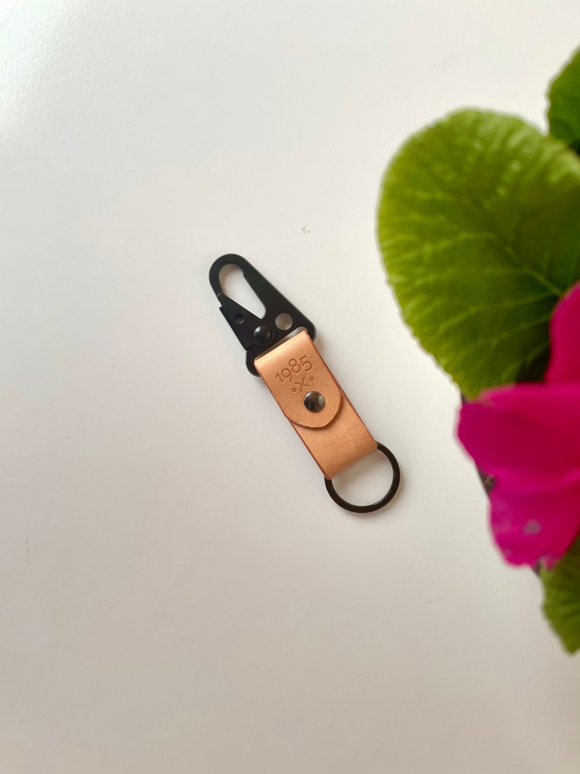 Carabiner keychain by 1985 Studio on a white table: a folded strap of natural vegetable-tanned leather stamped with the 1985 mark, between a black carabiner clip and a black split ring, a pink primrose beside it