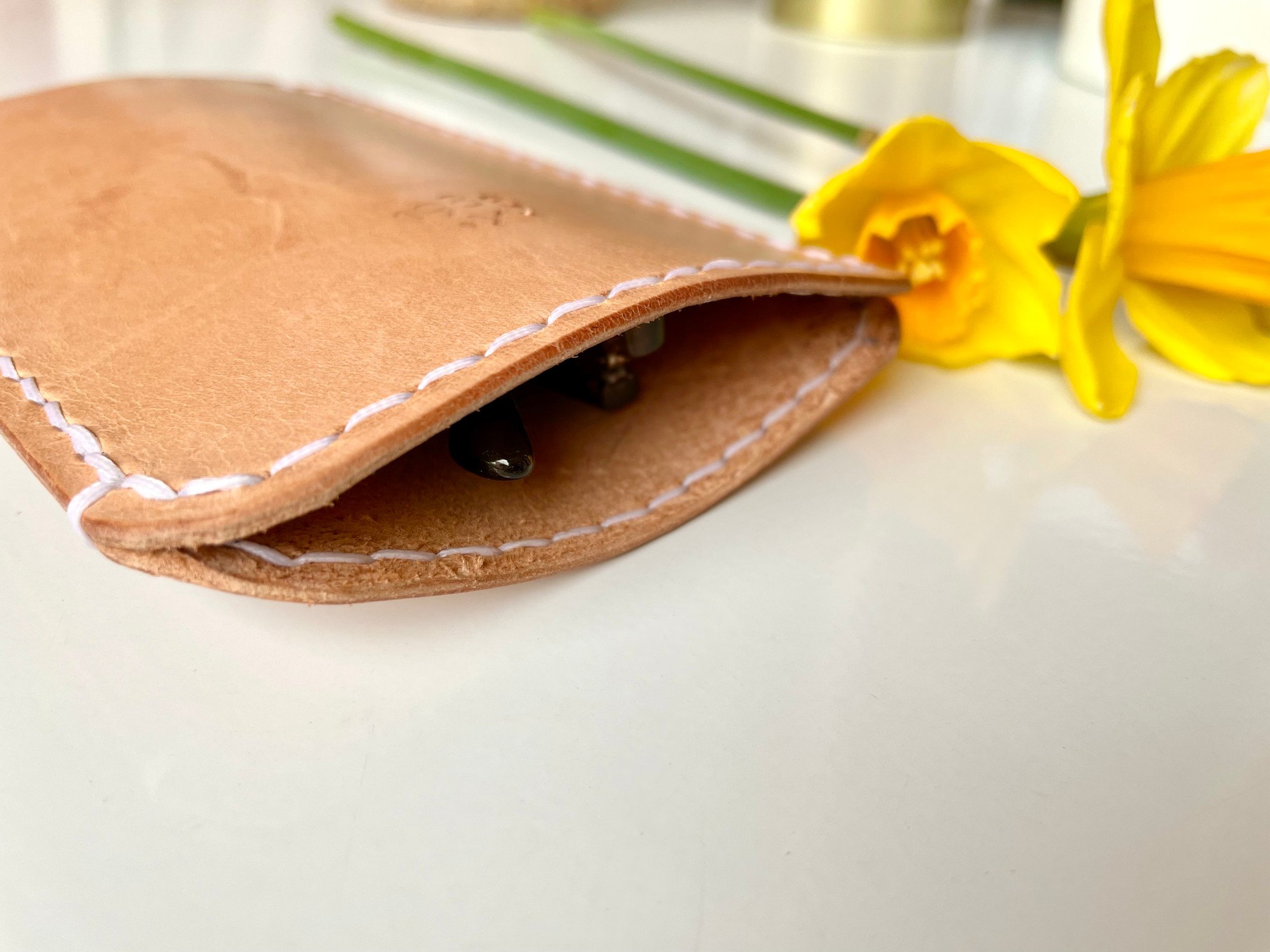 The opening of a slim sunglasses pouch by 1985 Studio up close, sunglasses inside and the pale saddle stitch running along the vegetable-tanned leather