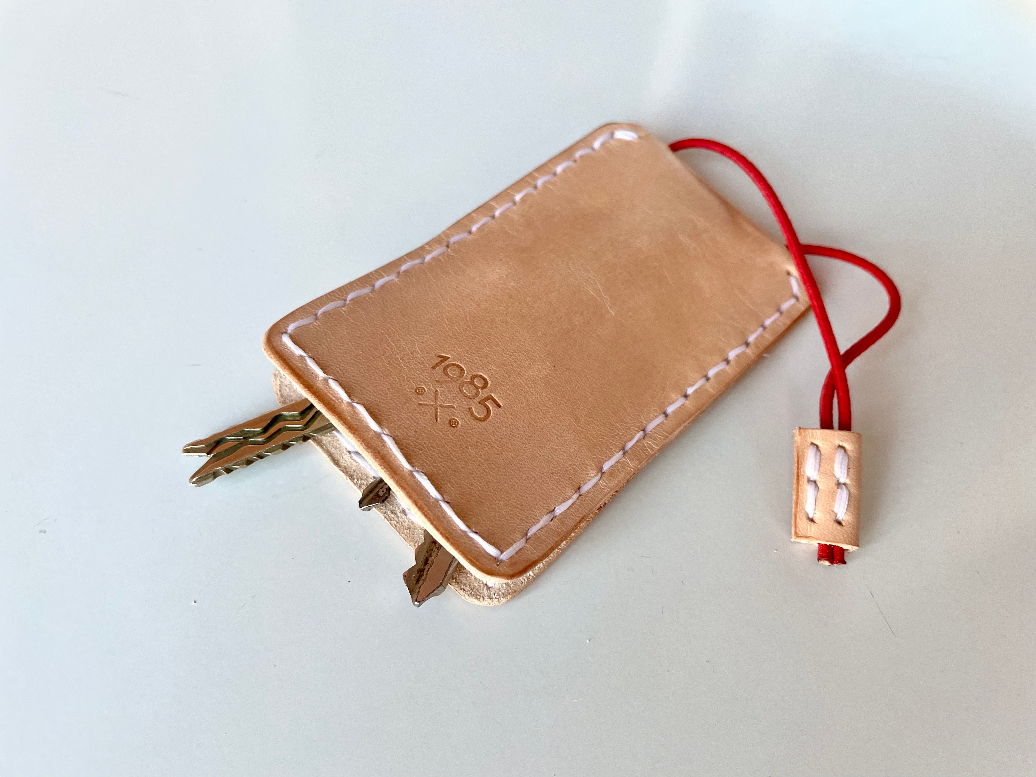 Slim key case by 1985 Studio in natural vegetable-tanned leather, stamped with the 1985 mark, keys peeking out of the bottom and a red elastic cord with a leather tab out of the top