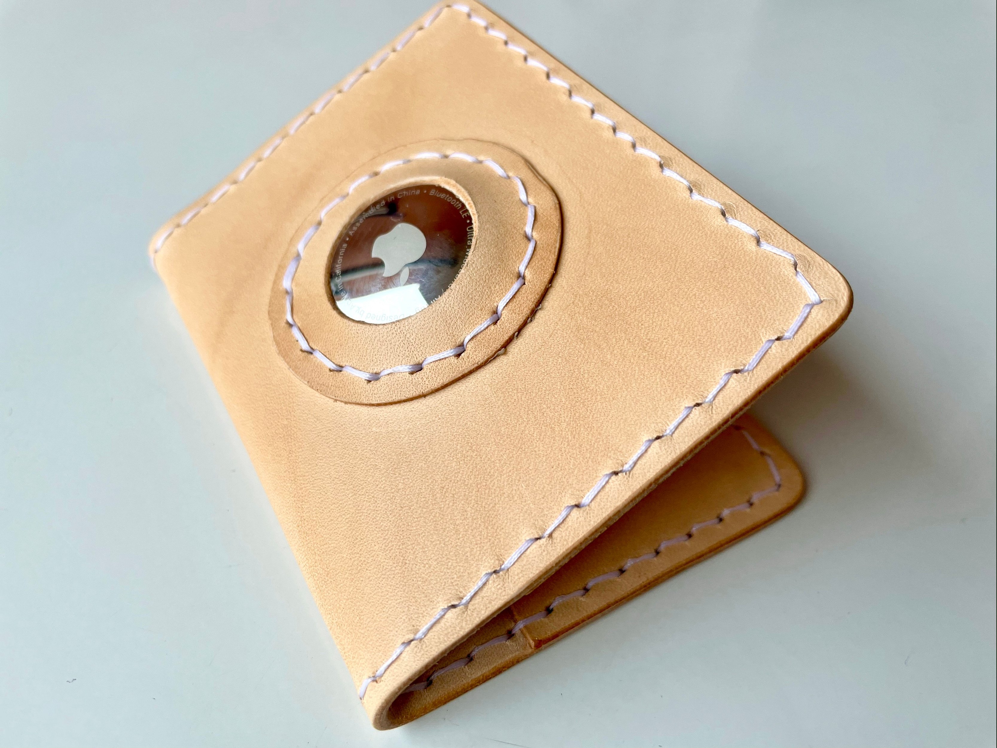 AirTag card wallet by 1985 Studio folded shut, natural vegetable-tanned leather stitched in white, an AirTag showing through the round window in its front