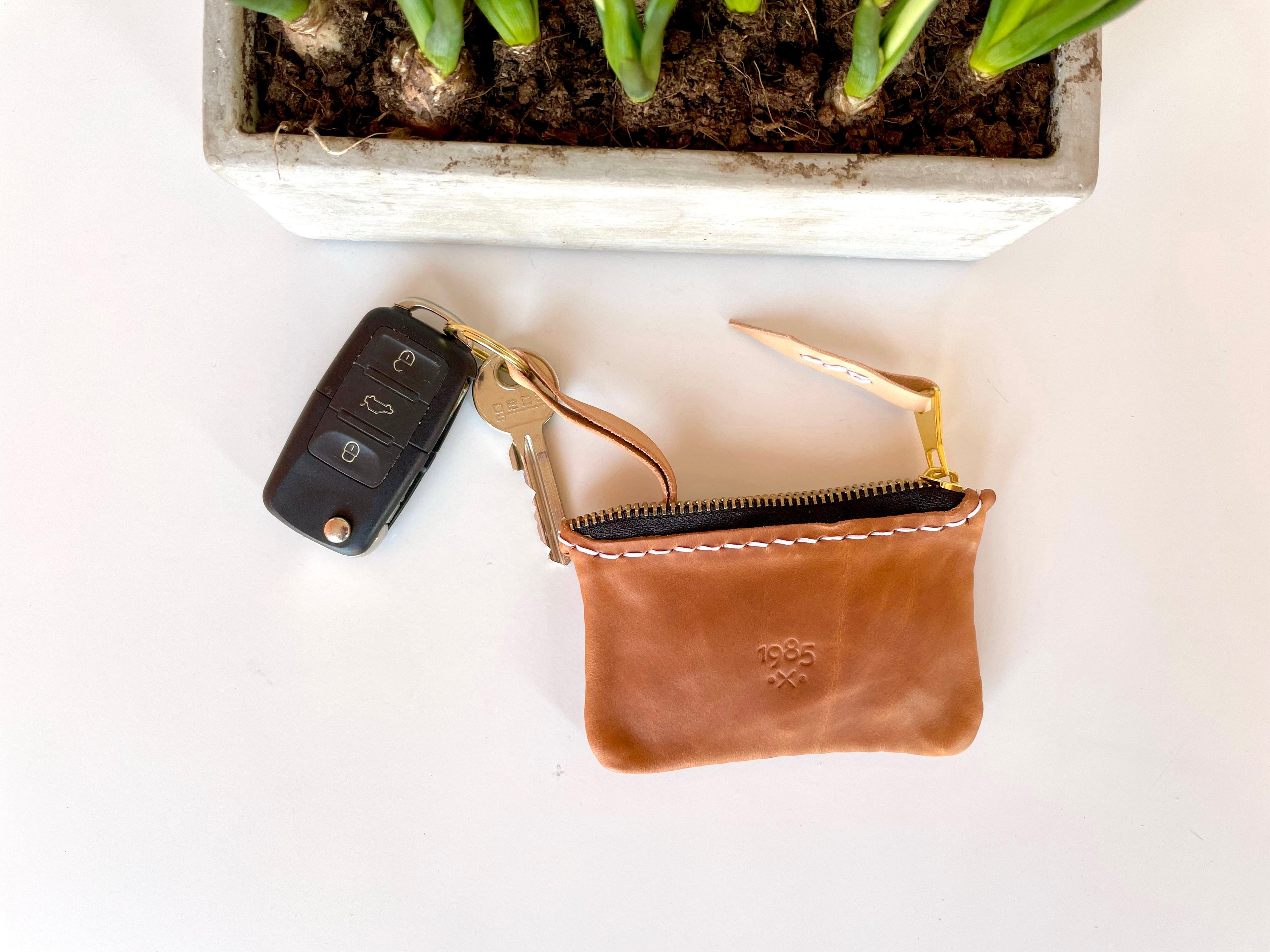 Car key pouch by 1985 Studio in vegetable-tanned leather on a white table, a car key and a house key on its brass ring, a planter of green shoots behind it