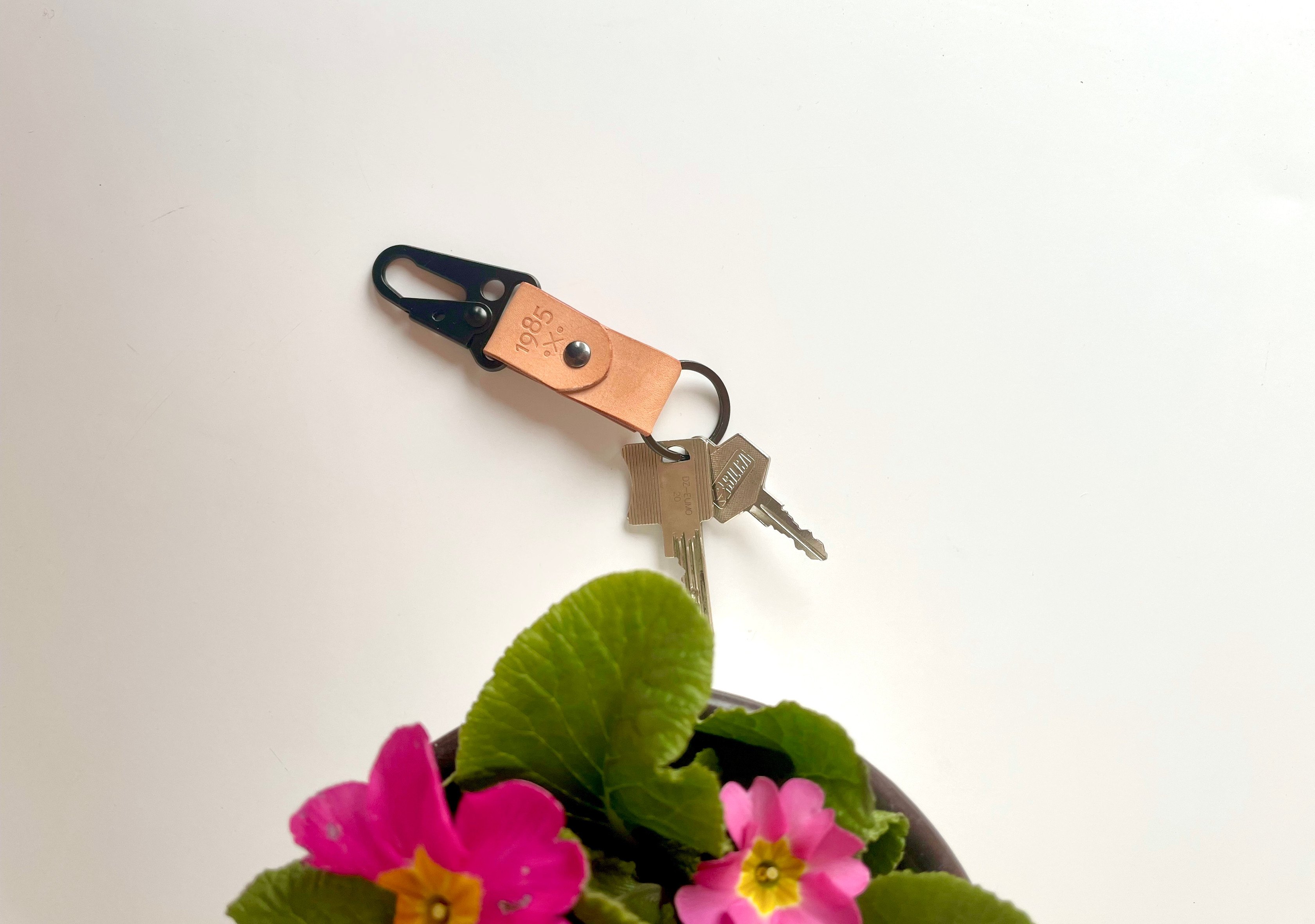 Carabiner keychain by 1985 Studio in vegetable-tanned leather holding two keys on a white table, pink primroses below it