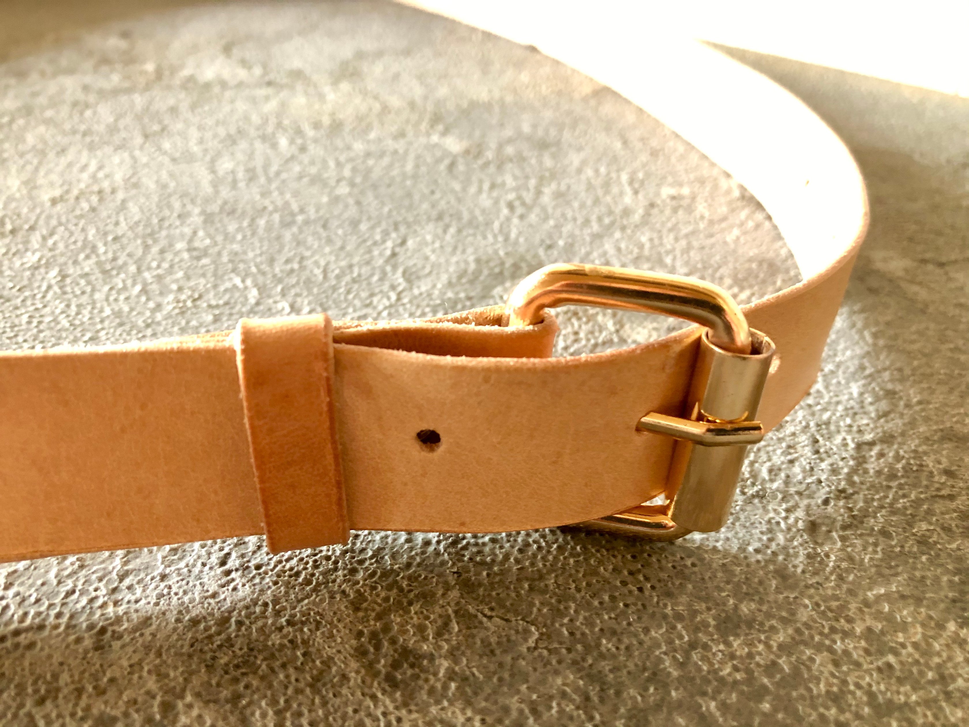 The brass roller buckle of a natural leather belt by 1985 Studio, the pale vegetable-tanned leather through its keeper, on grey stone