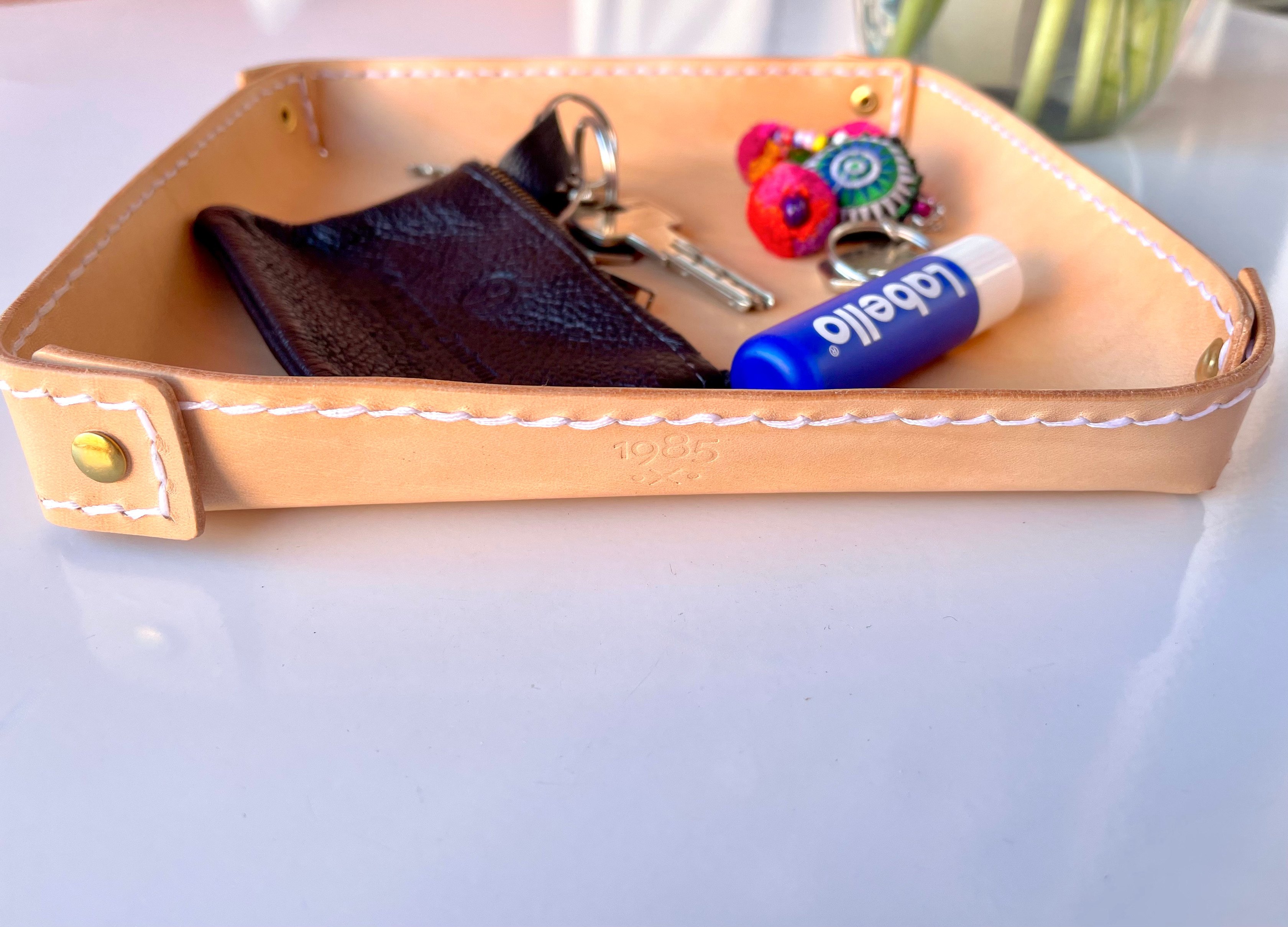 Fold-over valet tray by 1985 Studio from the side: its stitched edge of vegetable-tanned leather, brass rivets at the corners and the 1985 mark stamped on the front, keys and a zip pouch inside
